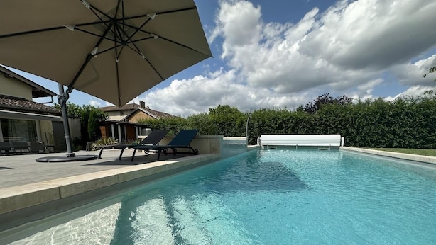 A swimming pool with a clear blue water, a lounge area with chairs and an umbrella, and a view of houses and trees in the background.