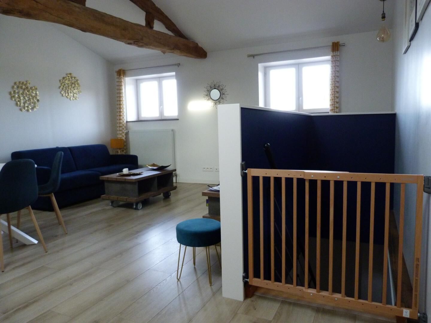 Een zithoek met een blauwe bank, een houten salontafel en een blauwe poef. Op de voorgrond is een houten leuning te zien.