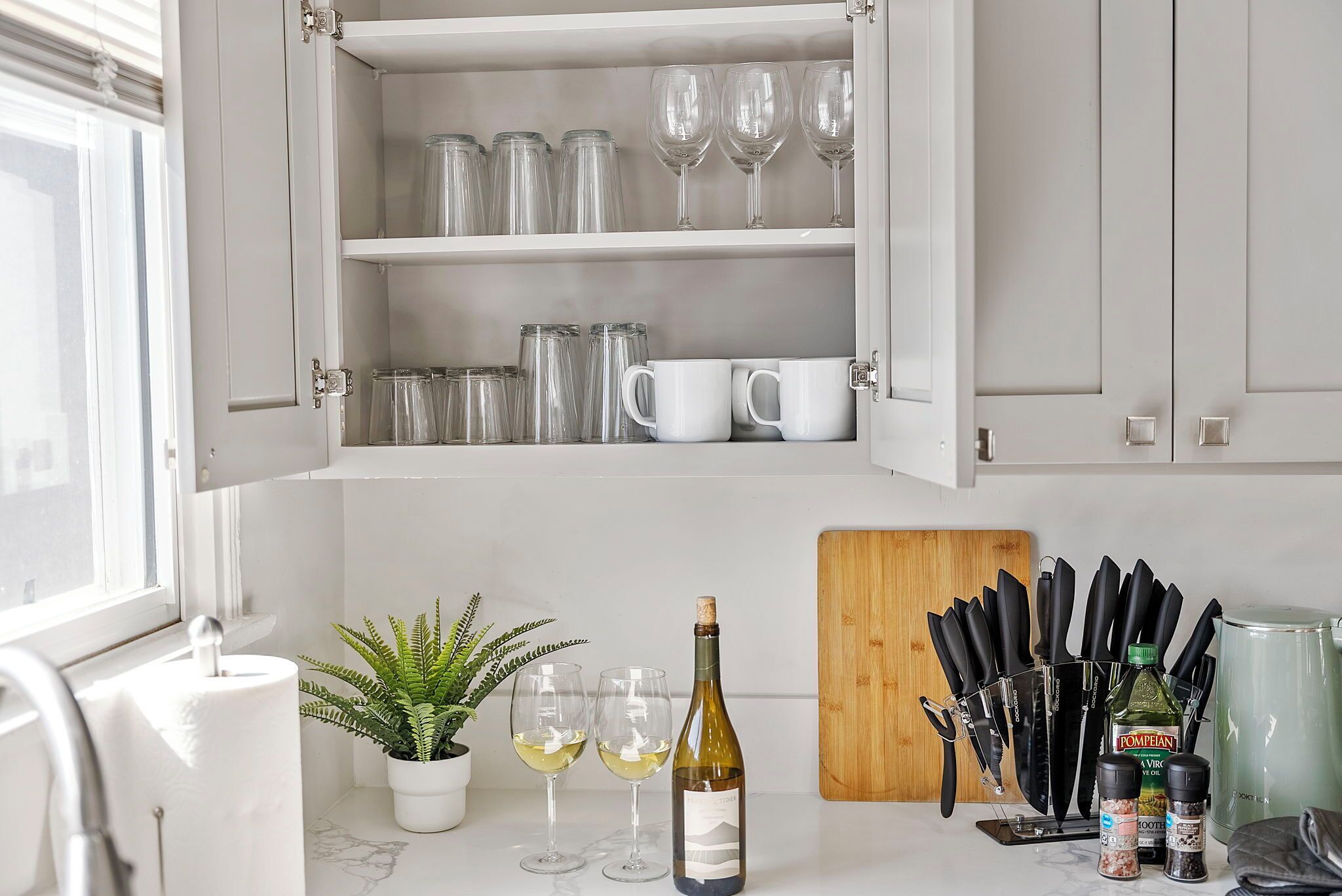 A kitchen with open cabinets containing wine glasses and mugs, a potted fern, a cork wine bottle, and a cutting board with knives.