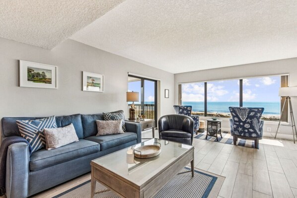 A modern living room with a blue sofa, a leather armchair, and a coffee table. There are large windows offering a view of the ocean.
