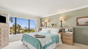 A bedroom with a large window, a bed, bedside tables, a dresser, and a painting on the wall.