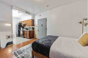 Une chambre à coucher de taille réduite, comprenant un lit, une lampe de chevet et donnant sur un coin cuisine.