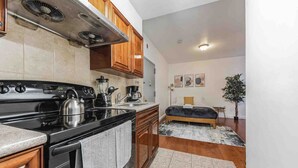 A compact kitchen with a stove, sink, and wooden cabinets. A bed with a dark headboard is visible in the adjacent room. - Studio Across the Street from Convention Center 2R (Philadelphia)