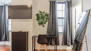A room with a wooden floor, a black dresser, a desk with a chair, a television mounted on the wall, and a window with grey curtains. - Studio Across the Street from Convention Center 2R (Philadelphia)