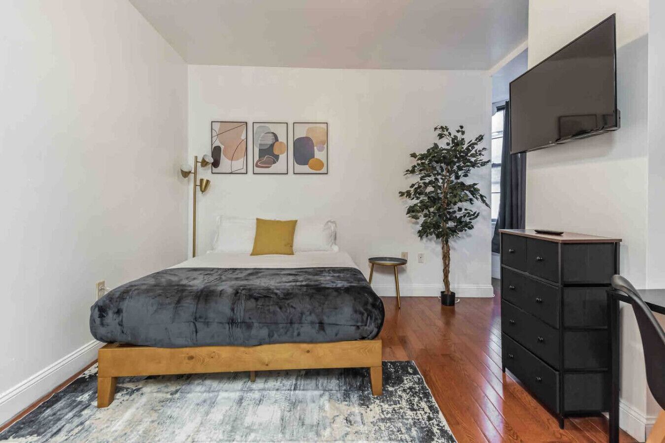 A bedroom with a bed, a dresser, a plant, and abstract art on the wall.