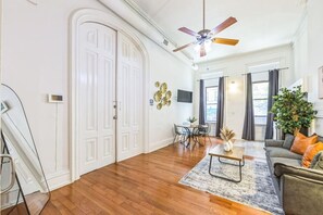 A spacious living room with hardwood flooring, a ceiling fan, a dining area with a table and chairs, and a sofa with cushions.