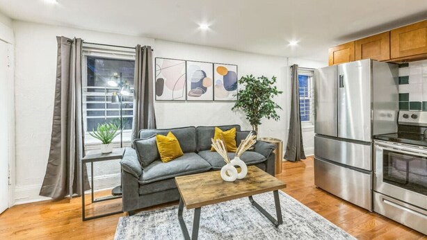 A modern kitchen with stainless steel appliances, a grey sofa with yellow cushions, and a wooden coffee table.