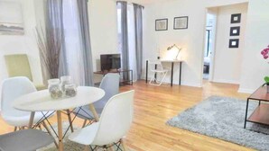 Un soggiorno moderno con pavimento in legno, un tappeto grigio, un tavolo da pranzo con sedie e una TV sullo sfondo.