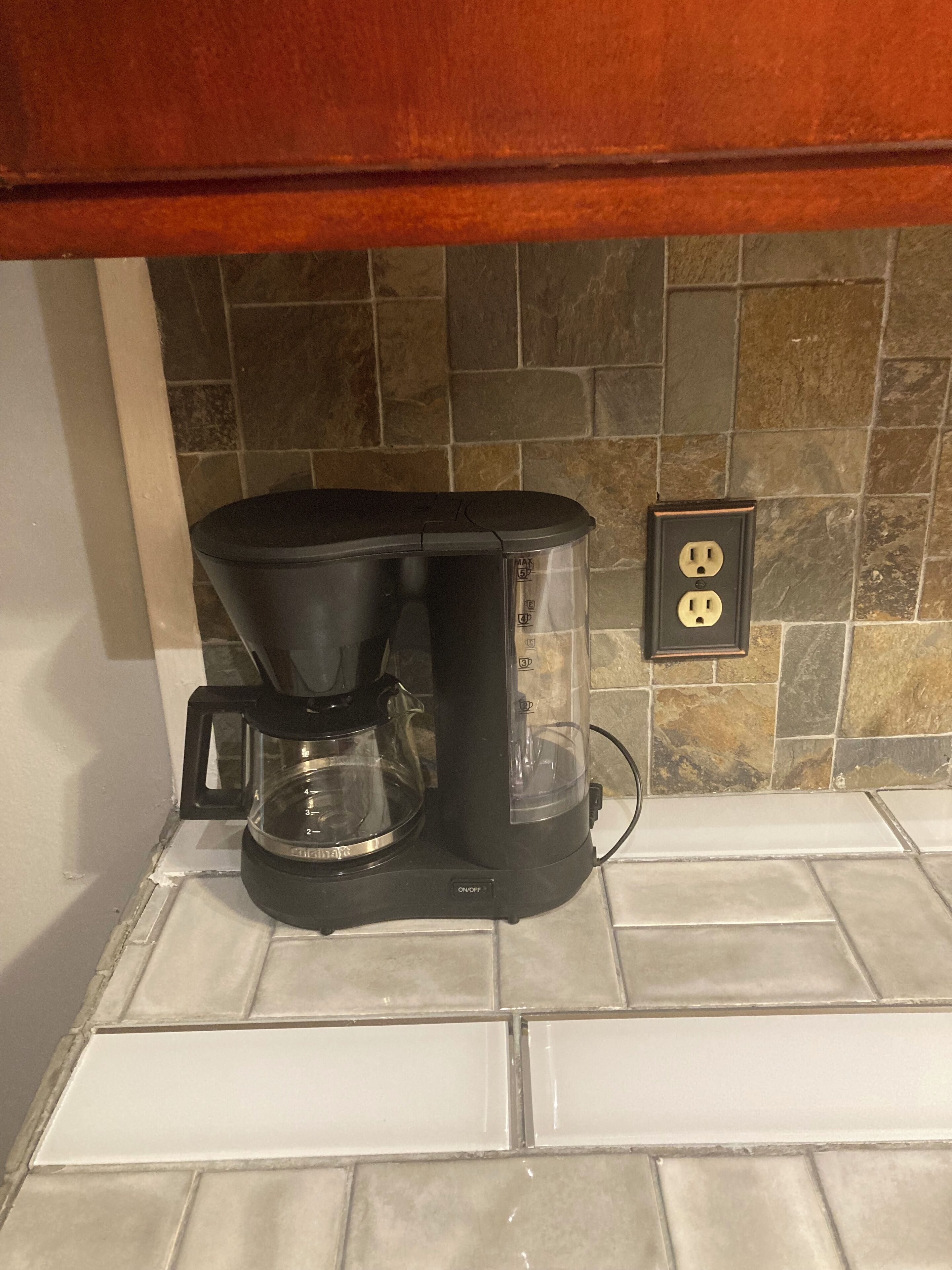 A coffee maker on a tiled kitchen counter with a stone backsplash and an electrical outlet.