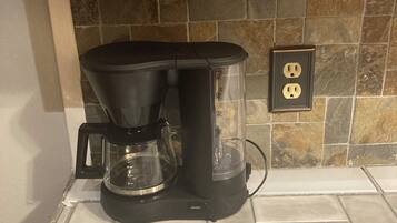 A coffee maker on a tiled kitchen counter with a stone backsplash and an electrical outlet.