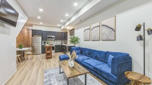 Uma sala de estar moderna com um sofá azul, uma mesa de centro de madeira e uma cozinha ao fundo.
