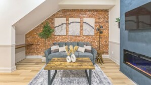 A cozy living room with a brick wall, a grey sofa, a wooden coffee table, and a fireplace. - New 2/2 Across from Fairmount Park | 10 mins to DT (Philadelphia)