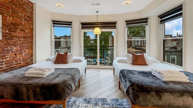 A modern bedroom with two beds, a brick wall, large windows, and a balcony. - New 2/2 Across from Fairmount Park | 10 mins to DT (Philadelphia)