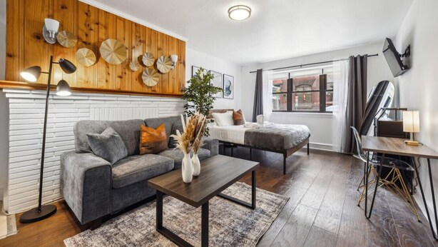 A modern living room with a grey sofa, a wooden accent wall, a dining area with a table and chairs, and a large window with curtains.