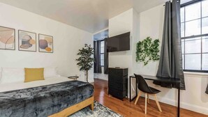 Un dormitorio con cama, escritorio, silla, televisor y ventana con cortinas.