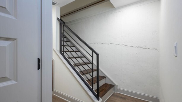 Una escalera con barandilla de metal negro y peldaños de madera, que conduce a un rellano con pared blanca y una puerta visible.