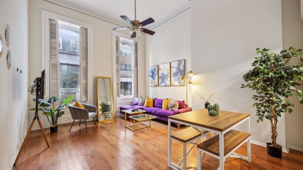A spacious living room with a wooden floor, a purple sofa, a coffee table, a ceiling fan, and a large window.