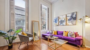 A living room with a purple sofa, a glass coffee table, a grey armchair, a tall mirror, and a floor lamp.