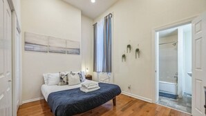 A bedroom with a wooden floor, a bed with a blue bedspread, white bedding, and a nightstand. There is a window with curtains, a wall-mounted plant, and a door leading to a bathroom.