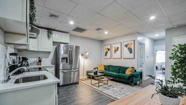 Eine moderne Küche mit einem Edelstahlkühlschrank, weißen Schränken und einer integrierten Spüle. Im Wohnbereich befindet sich ein grünes Sofa, ein Couchtisch und eine Pflanze in einem weißen Topf.