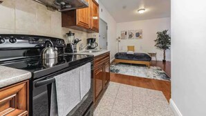 A compact kitchen with a stove, coffee maker, and cabinets. A bed is visible in the adjacent room with a rug and a plant.