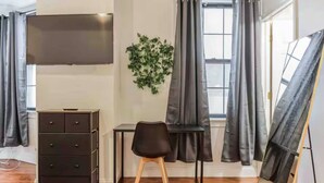A room with a wooden floor, a black desk, a black chair, a dresser, a TV mounted on the wall, a window with curtains, and a painting on an easel.