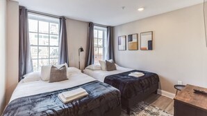 A modern hotel room with two beds, a desk, and large windows offering a city view.