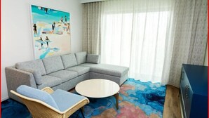 A modern living room with a grey sectional sofa, a wicker chair, a round coffee table, and a painting of beachgoers.