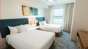 A hotel room with two beds, a large window with curtains, a ceiling fan, and a painting on the wall.