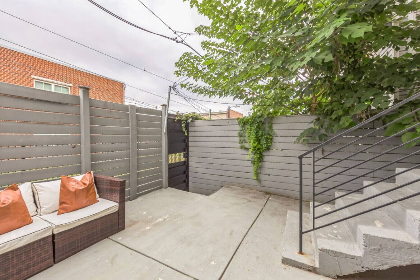 A backyard with a grey fence, a concrete patio, a metal staircase, and a brick bench with cushions.