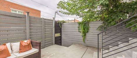 A backyard with a grey fence, a concrete patio, a metal staircase, and a brick bench with cushions.