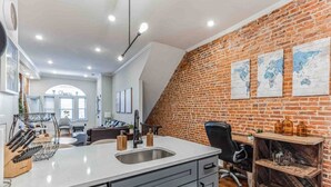 A modern kitchen with a large island, a brick wall, and a dining area in the background. - Large Bi-Level 3/2 w/Patio | 10 min to DT (Philadelphia)