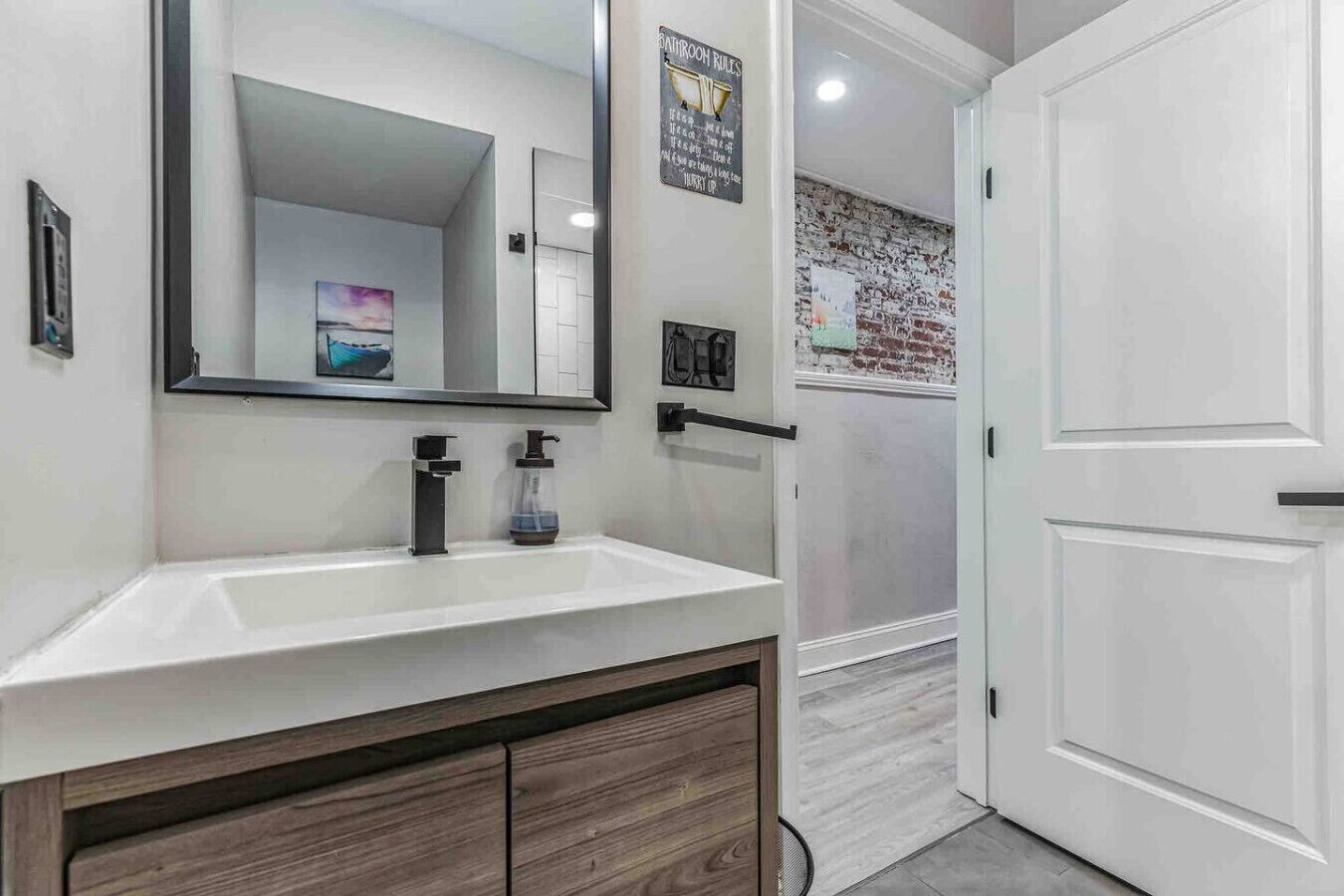 Une salle de bain moderne avec un lavabo blanc, un robinet noir et un grand miroir. On y trouve un meuble en bois sous le lavabo et une porte blanche sur la droite.