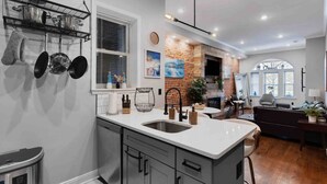 A modern kitchen with a large island, a brick wall, and a dining area with a couch and table. - Large Bi-Level 3/2 w/Patio | 10 min to DT (Philadelphia)