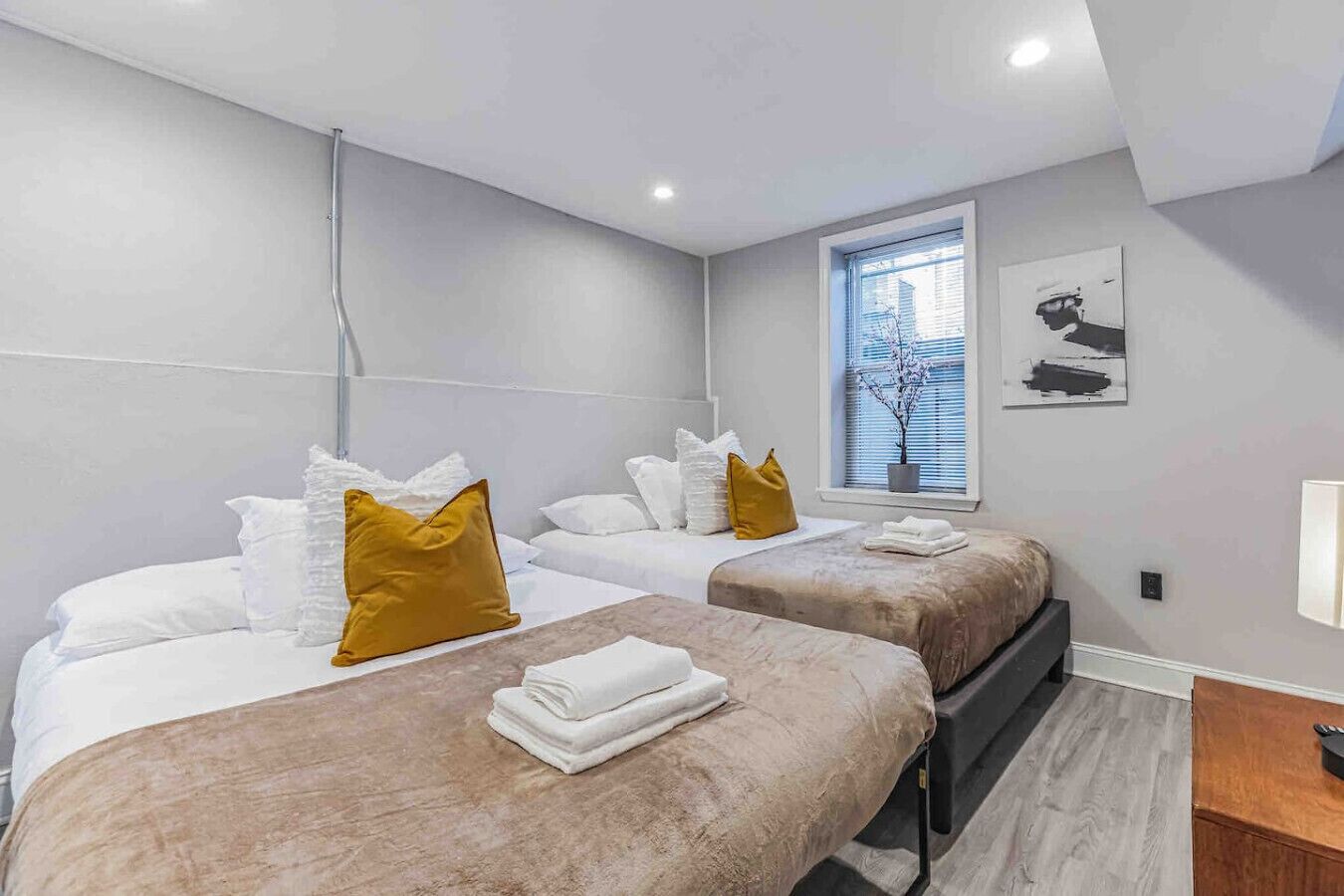 A hotel room with two beds, each with white bedding and a brown headboard. There is a window with blinds, a small potted plant, and a wall-mounted light fixture.