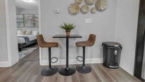A modern kitchen with a dining area, featuring a black table, two wooden bar stools, a potted plant, and decorative wall art.