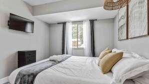 A bedroom with a bed, a TV mounted on the wall, a window with curtains, and a small dresser.