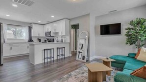 A modern kitchen with white cabinets and a central island, a living area with a green sofa and a flat-screen TV, and a wooden coffee table.
