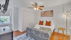 A bedroom with a bed, a ceiling fan, a desk, a window, and artwork on the wall.