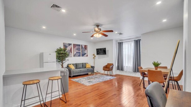 A spacious living area with a wooden floor, a grey sofa, a dining table with chairs, a ceiling fan, and a TV mounted on the wall.