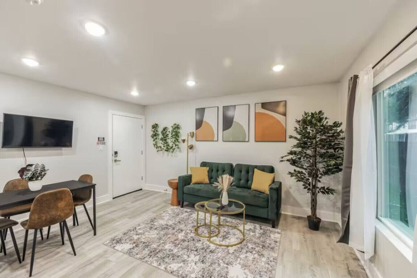 Ein modernes Wohnzimmer mit einem grünen Sofa, einem Esstisch mit Stühlen, einem an der Wand montierten Fernseher und einem großen Fenster.