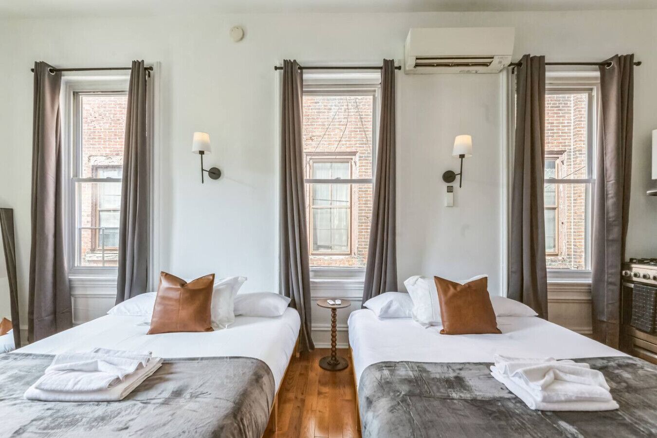 A hotel room with two beds, a small table, and a view of a brick building through large windows.