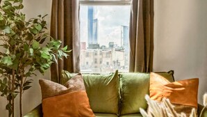 Habitació amb un sofà verd, una planta en testos i vista a edificis per la finestra.