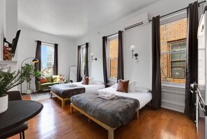 A modern hotel room with two beds, a small seating area, a dining table, and large windows with curtains. - New Studio in Center City | Sleeps 4 | Fast Wifi (Philadelphia)