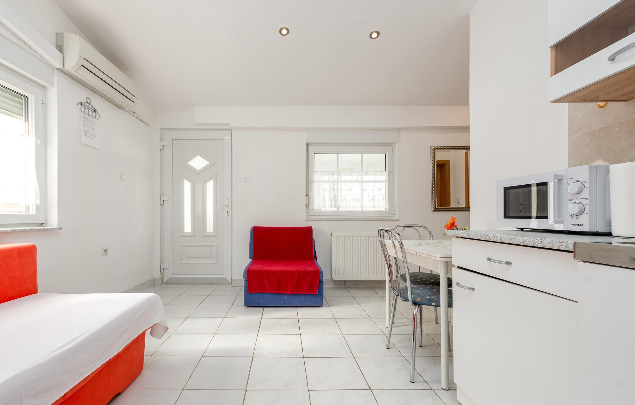 A compact hotel room with a bed, a red chair, a dining table with chairs, a microwave, and a small kitchenette.