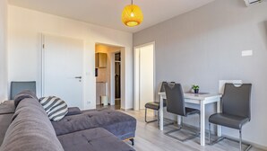 Ein modernes Wohnzimmer mit einem grauen Sofa, einem Esstisch mit Stühlen und einem durchgehenden Gang sichtbaren Küchenbereich.