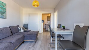 Ein modernes Wohnzimmer mit einem grauen Ecksofa, einem Esstisch mit zwei Stühlen und einer Küche im Hintergrund.