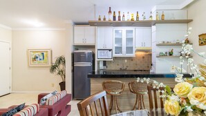 Una cocina moderna con armarios blancos, microondas integrado y refrigerador de acero inoxidable. Hay un comedor con mesa y sillas, y una sala de estar con sofá y una planta.