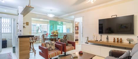 A modern living room with a sofa, a coffee table, and a TV. There is a kitchen area with a dining table and chairs.
