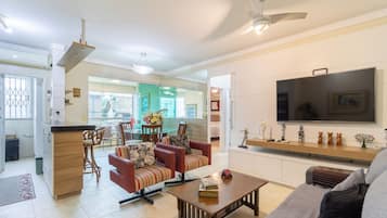 A modern living room with a sofa, a coffee table, and a TV. There is a kitchen area with a dining table and chairs.